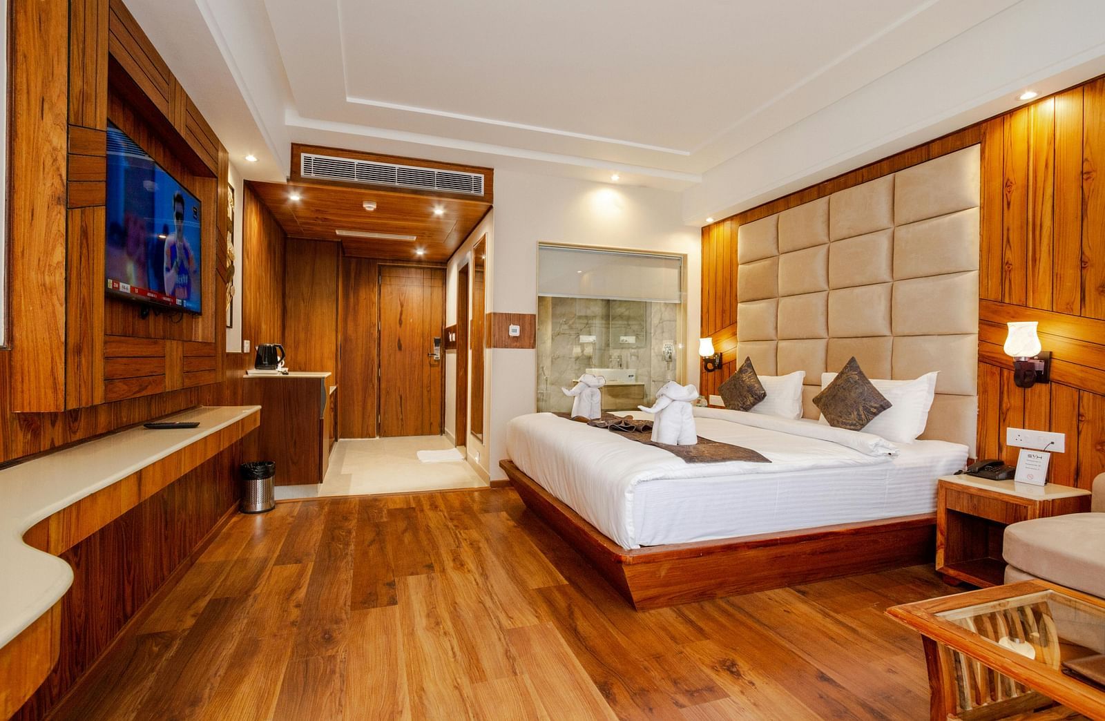 Spacious hotel room with modern furnishings, a large comfortable bed, wall-mounted TV, wooden flooring, and a glass-enclosed bathroom area.