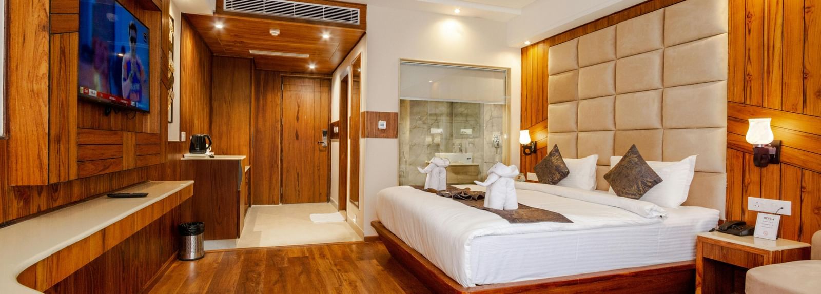 Spacious hotel room with modern furnishings, a large comfortable bed, wall-mounted TV, wooden flooring, and a glass-enclosed bathroom area.