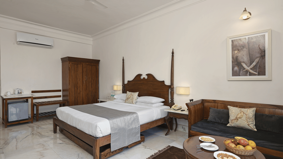 Deluxe Room with Balcony at Fateh Safari Resort, Kumbhalgarh featuring a bed, a wardrobe, an air-conditioner, a cabinet table, a sofa with centre table, and bedside table on either side of the bed.