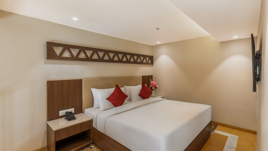 Bedroom at EsthereaRaj Leela, Ranakpur, featuring a king size bed, red accent pillows, a decorative wall panel, and a bedside table.