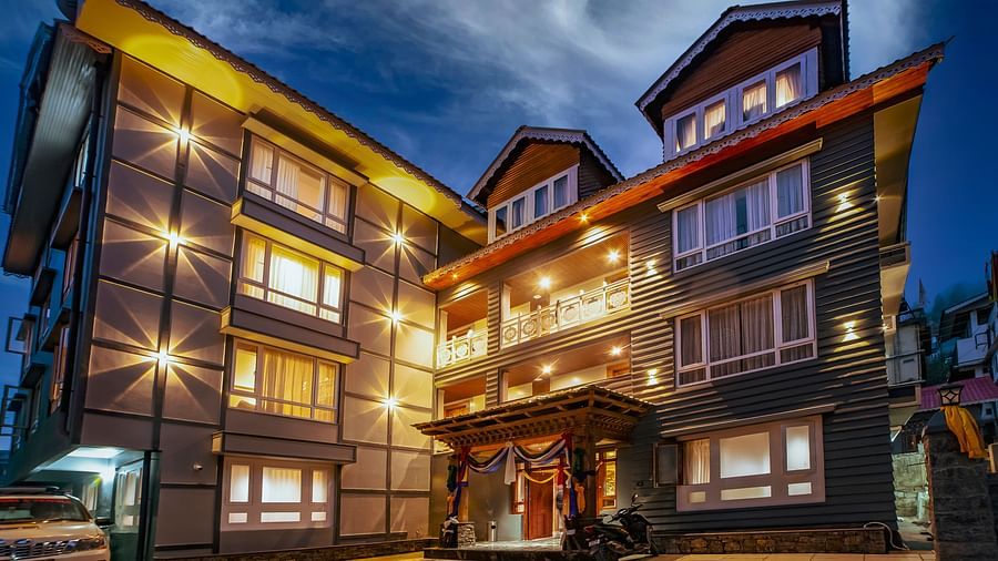 Modern, multi-story hotel building illuminated by warm exterior lights at dusk, featuring large windows, a welcoming entrance with decorative drapes, and a parked vehicle in the driveway - Yashshree Lachen.