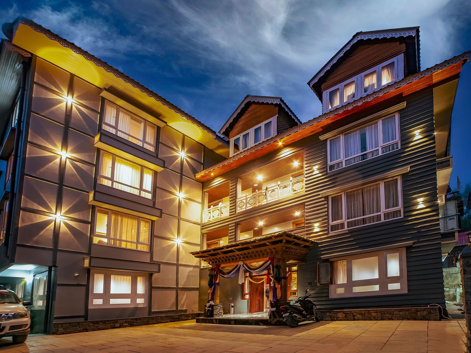 Modern, multi-story hotel building illuminated by warm exterior lights at dusk, featuring large windows, a welcoming entrance with decorative drapes, and a parked vehicle in the driveway - Yashshree Lachen.
