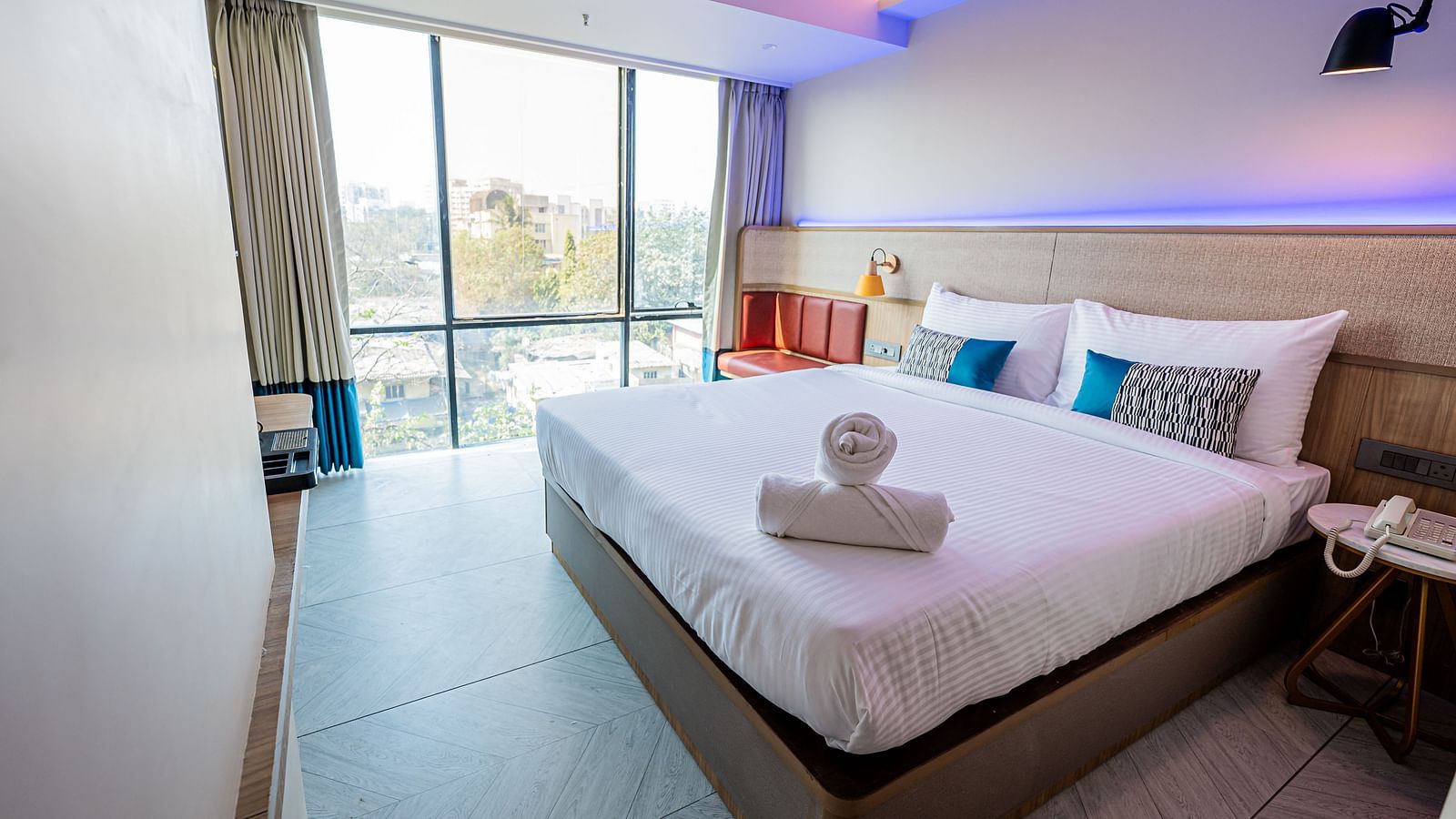 A spacious room at Indie Stays Mumbai International Airport features a large bed with neatly rolled towels, a window with the curtains open, and the room lit with cool lighting.