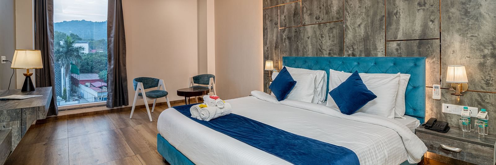 An image of a premium room with a bed, bed side tables, lamps, two chairs, a coffee table, and an expansive glass window overlooking the view - DLS Ganga Bliss, Haridwar
