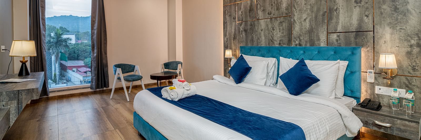An image of a premium room with a bed, bed side tables, lamps, two chairs, a coffee table, and an expansive glass window overlooking the view - DLS Ganga Bliss, Haridwar