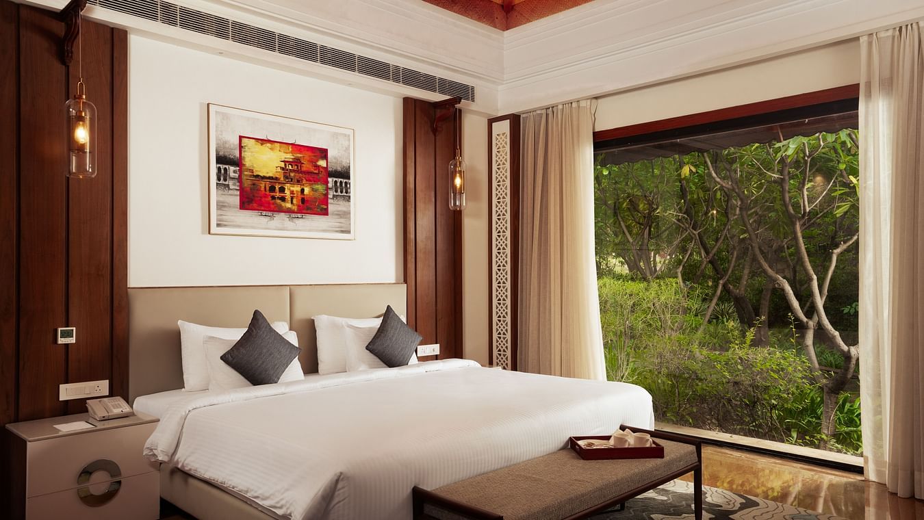 A spacious and well-lit bedroom at The Ananta Udaipur, featuring a king-sized bed, a wooden headboard, and a balcony overlooking the lush greenery.
