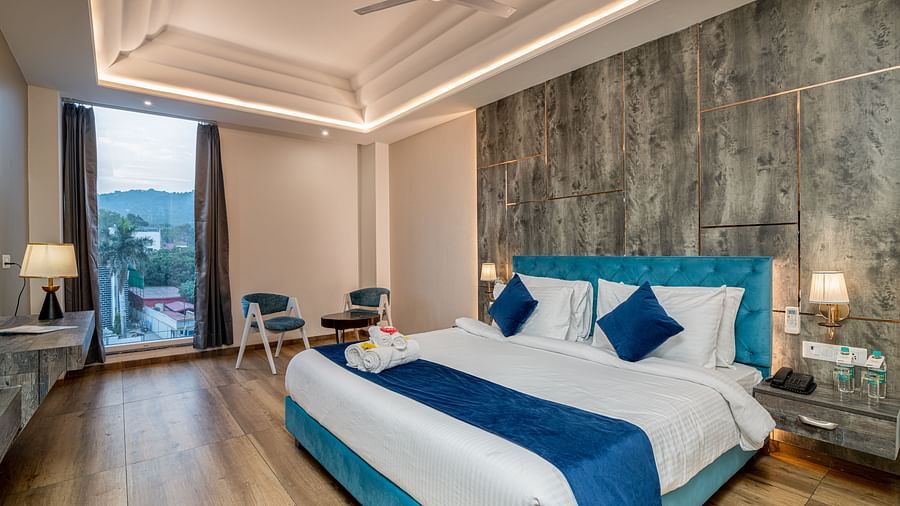 An image of a premium room with a bed, bed side tables, lamps, two chairs, a coffee table, and an expansive glass window overlooking the view - DLS Ganga Bliss, Haridwar