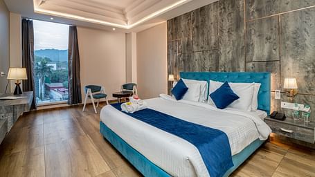 An image of a premium room with a bed, bed side tables, lamps, two chairs, a coffee table, and an expansive glass window overlooking the view - DLS Ganga Bliss, Haridwar