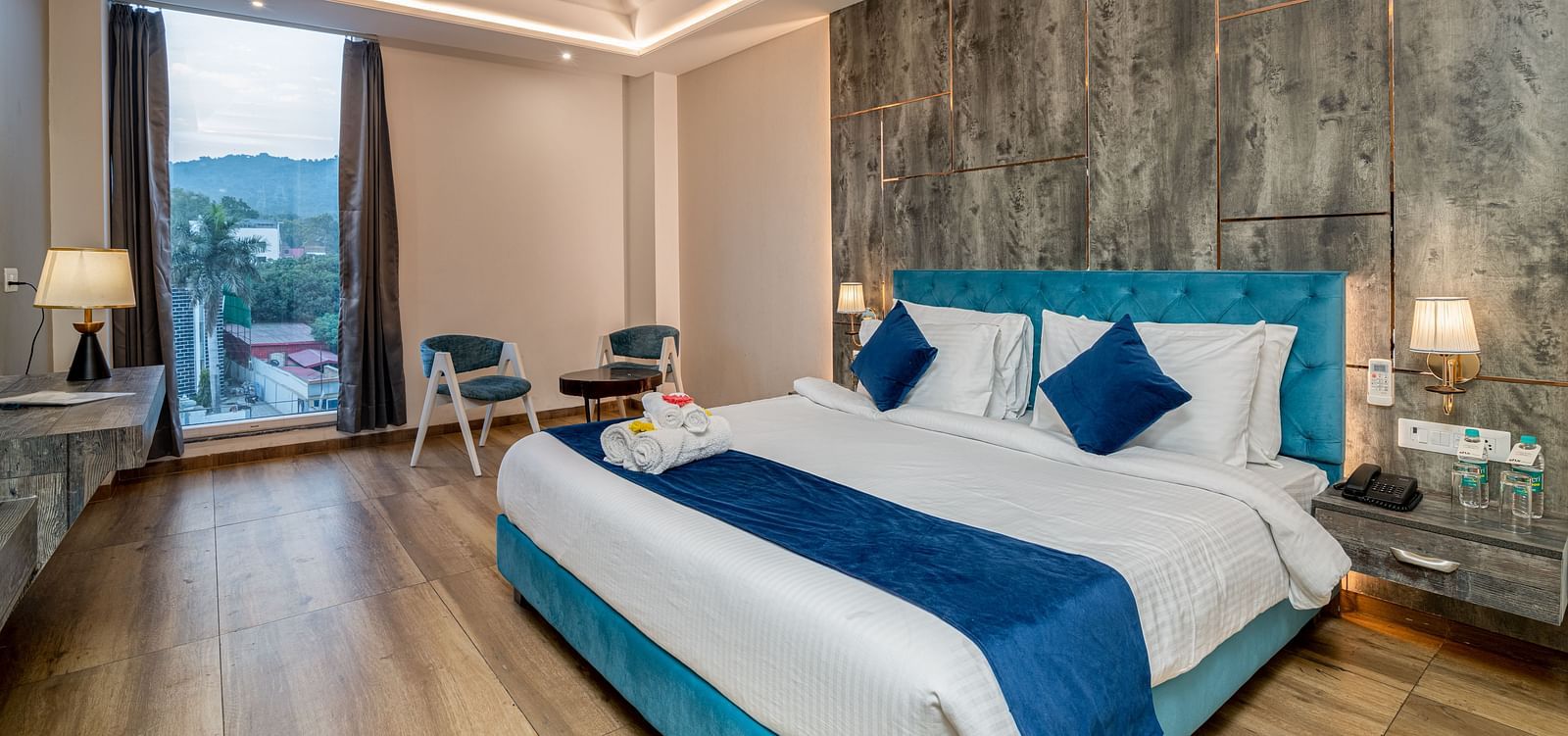 An image of a premium room with a bed, bed side tables, lamps, two chairs, a coffee table, and an expansive glass window overlooking the view - DLS Ganga Bliss, Haridwar