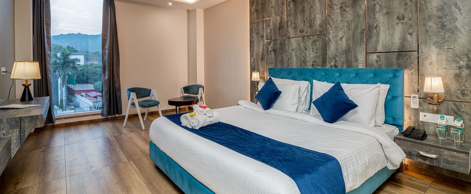 An image of a premium room with a bed, bed side tables, lamps, two chairs, a coffee table, and an expansive glass window overlooking the view - DLS Ganga Bliss, Haridwar