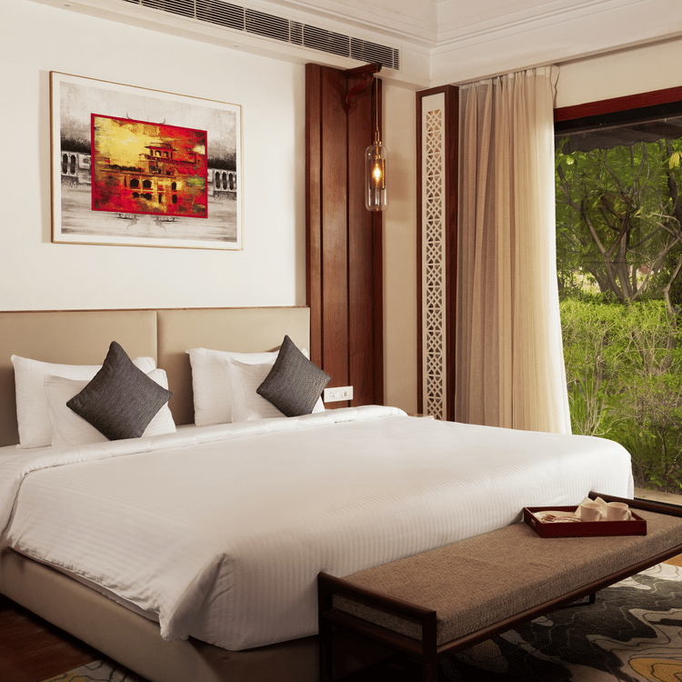 A spacious and well-lit bedroom at The Ananta Udaipur, featuring a king-sized bed, a wooden headboard, and a balcony overlooking the lush greenery.