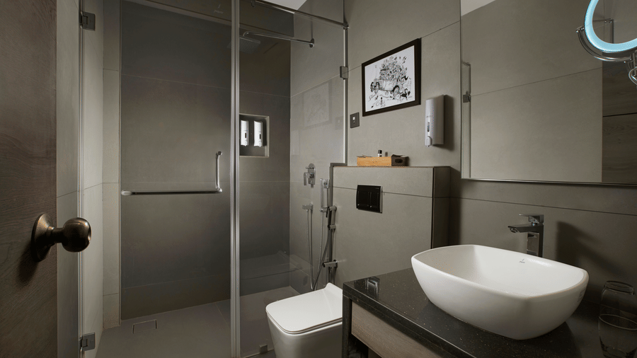 Modern bathroom at Ocean Palms Goa featuring a glass-enclosed shower, vessel sink, toilet, grey stone wall tiles, and framed artwork above the vanity.
