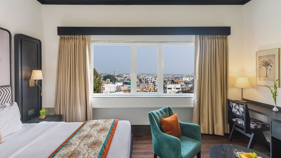 a spacious room featuring a king size bed, one comfortable green armchair, a cabinet with a table lamp and a window overlooking the city - Hotel Rajdarshan, Udaipur.