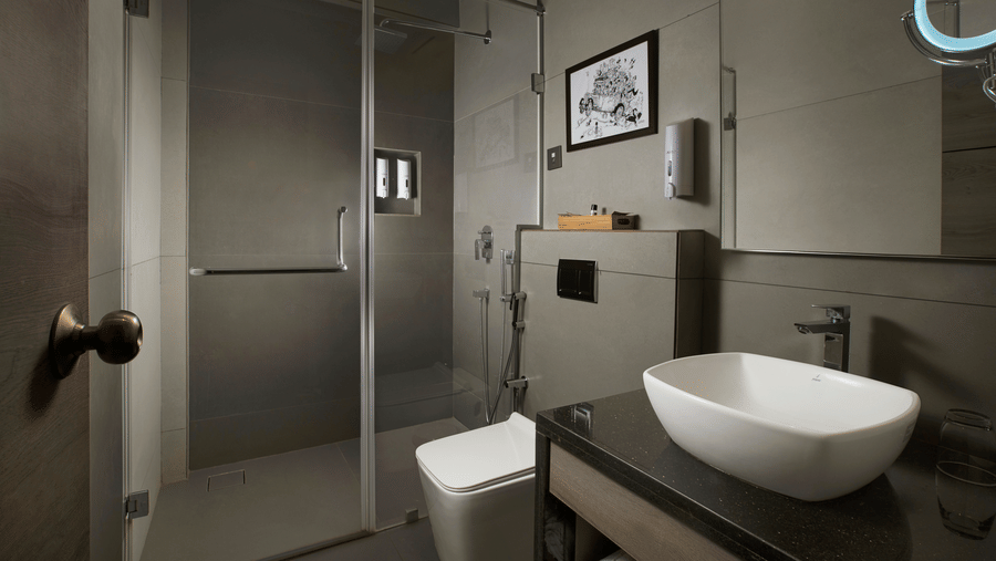 Modern bathroom at Ocean Palms Goa featuring a glass-enclosed shower, vessel sink, toilet, grey stone wall tiles, and framed artwork above the vanity.