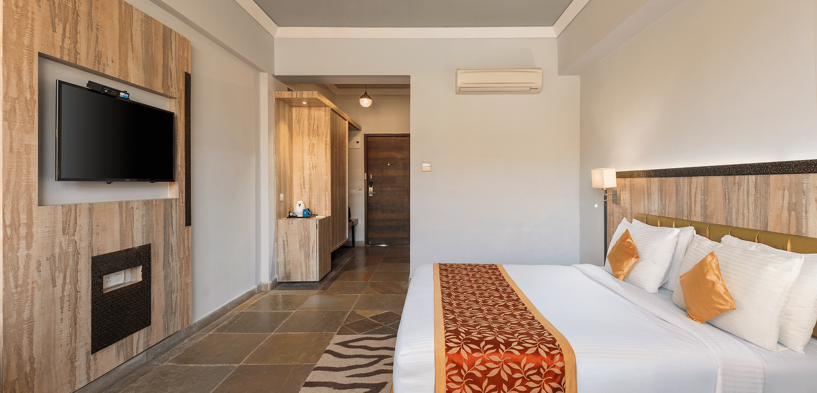 A hotel room with a bed, TV, door leading to another room, and a patterned rug on the floor - Via Lakhela Resort & Spa, Kumbhalgarh