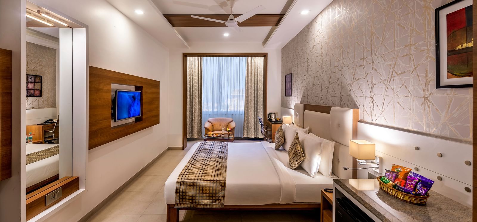 A room with a king size bed, a seating area by a balcony, a work desk, and a wall-mounted TV.