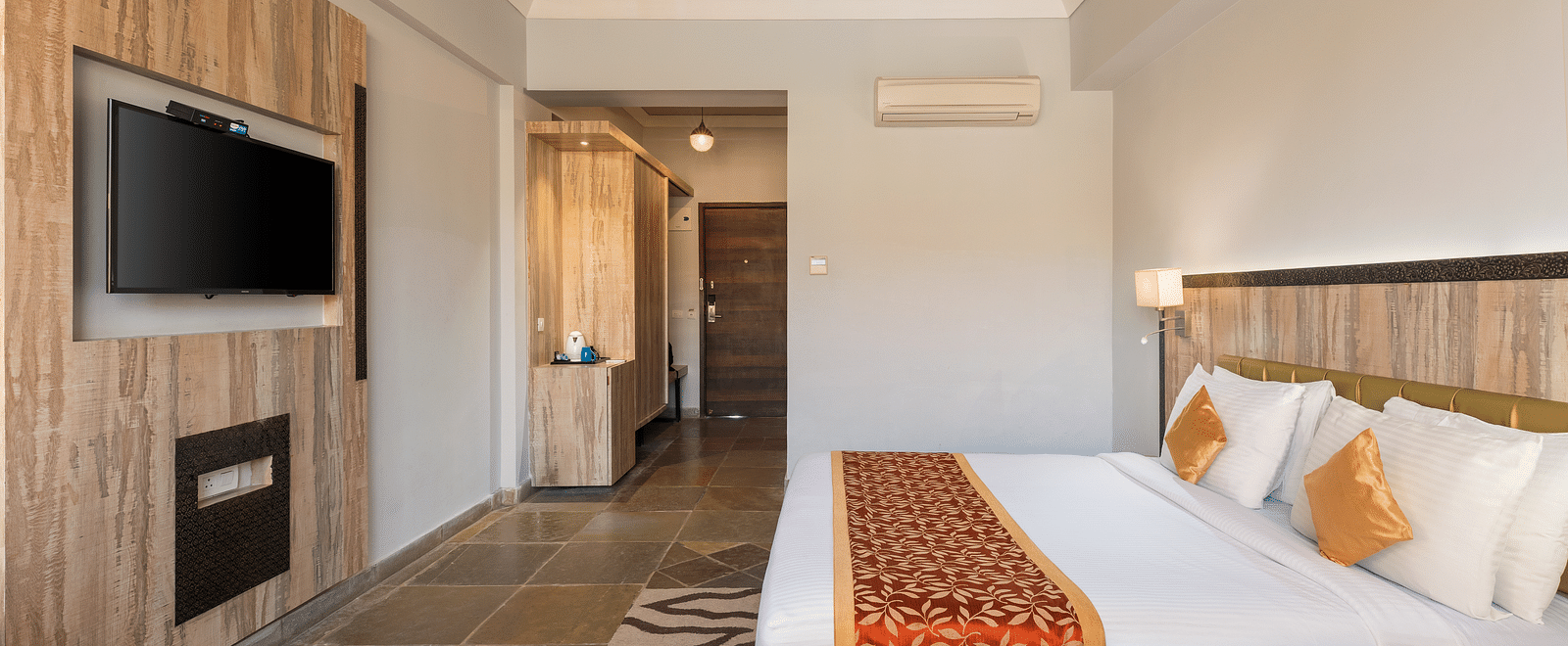 A hotel room with a bed, TV, a door leading to another room, and a patterned rug on the floor - Via Lakhela Resort & Spa, Kumbhalgarh