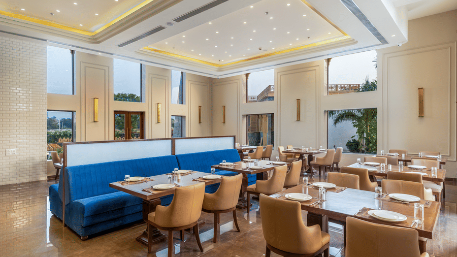 The restaurant at EsthereaRaj Leela, Ranakpur, featuring large windows, set tables, and a central blue upholstered booth partition.