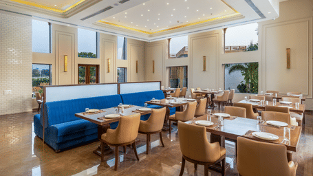 The restaurant at EsthereaRaj Leela, Ranakpur, featuring large windows, set tables, and a central blue upholstered booth partition.