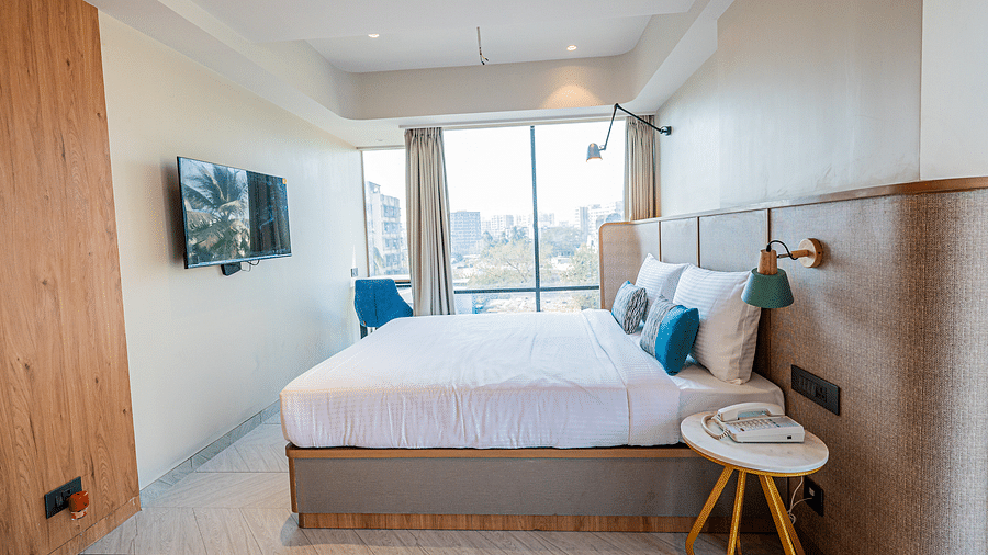 A side view of a spacious room at Indie Stays Mumbai International Airport, featuring a large bed, a three-legged bedside table, a wall-mounted TV, and a wide window with curtains drawn aside.