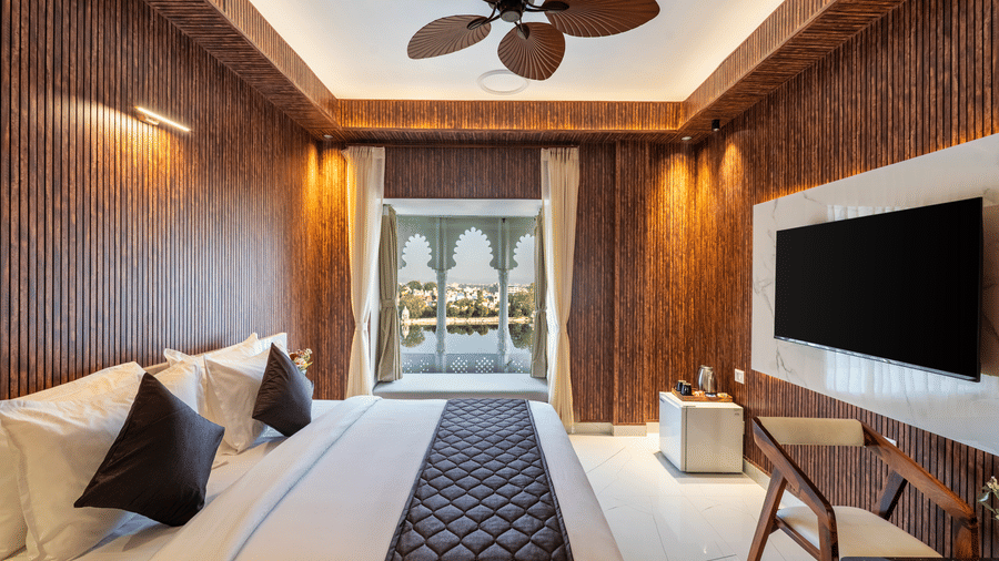 Interior of a contemporary hotel room featuring a bed, wooden cladding, a wicker fan, and an outside view through the large window.