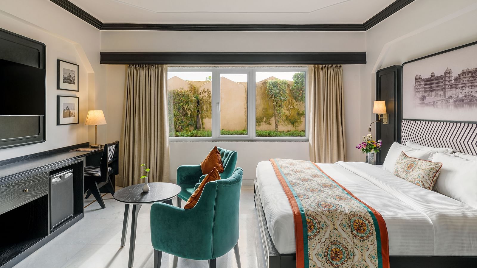 a spacious room featuring a king size bed, two comfortable green armchairs, with a table lamps and a window - Hotel Rajdarshan, Udaipur.