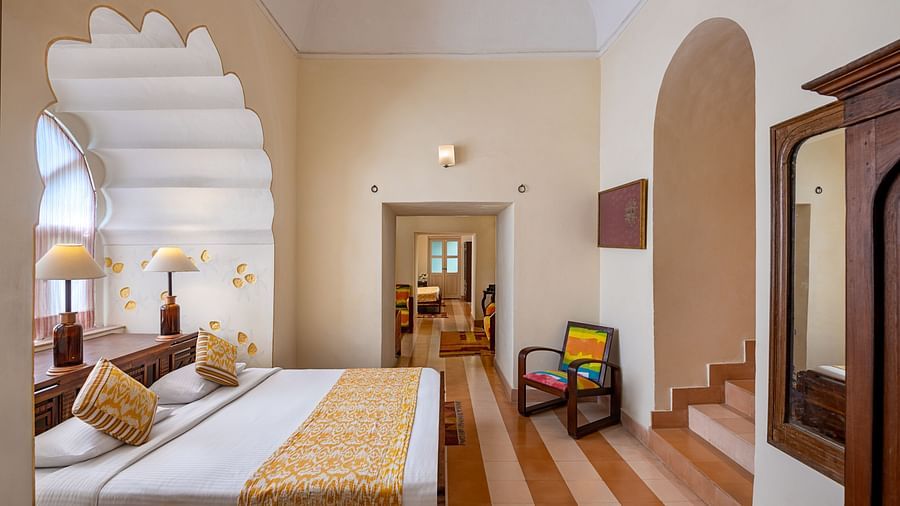 Room with bed, patterned pillows and runner, arched alcove, striped floor, furniture including chair and wardrobe, and doorway leading to another room - Tijara Fort-Palace - 19th Century, Alwar.
