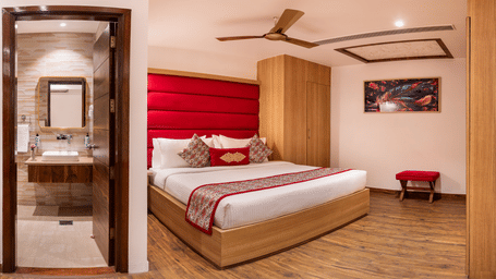 View of a Standard Room at Lords Inn Amritsar with a king bed, red headboard, wooden flooring, ceiling fan, and an attached bathroom beyond.