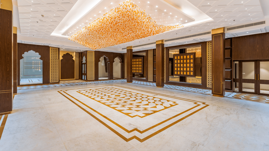 A wide, banquet hall at EsthereaRaj Leela, Ranakpur, featuring a large rectangular crystal chandelier and patterned marble flooring.