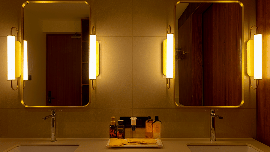 Bathroom vanity area at SUKAVI with 2 sinks, 2 rectangular mirrors with vertical lights, and toiletries placed on the counter.