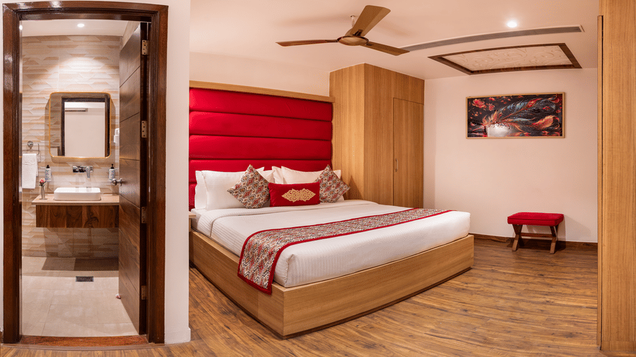 View of a Standard Room at Lords Inn Amritsar with a king bed, red headboard, wooden flooring, ceiling fan, and an attached bathroom beyond.