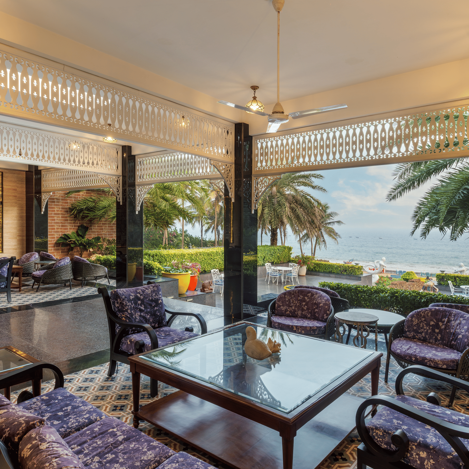 Sea-facing lounge with purple cane chairs, glass tables, white fretwork arches and a patterned tiled floor at MAYFAIR Palm Beach Resort, Gopalpur-on-Sea.