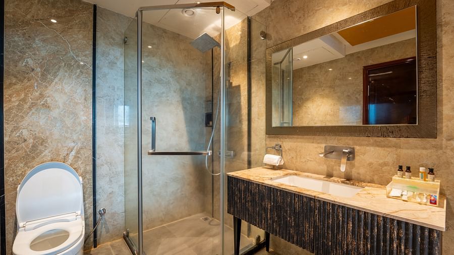 Executive Room bathroom features a rectangular mirror above the wash basin, a shower head to the right, a glass panel separating the shower area, and a western toilet at Crescent Spa & Resort, Indore