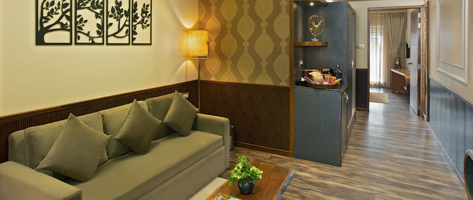Image of Coral Suite at Coral Reef Hotel and Spa in Port Blair featuring a three-seater sofa with a wooden coffee table and a lamp in the corner as the shelf displays showpieces and snacks and the door leads to the bedroom