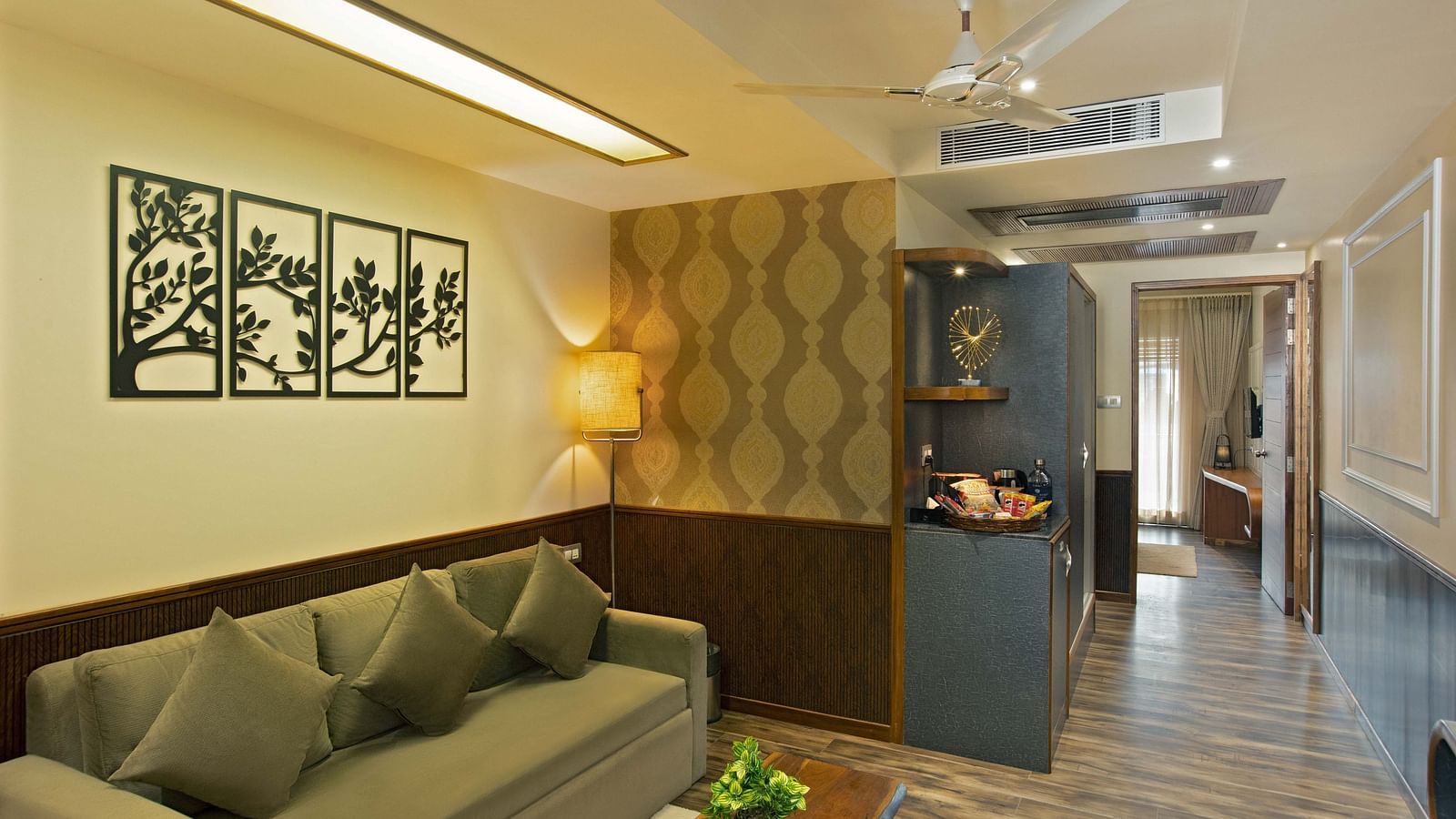 Image of Coral Suite at Coral Reef Hotel and Spa in Port Blair featuring a three-seater sofa with a wooden coffee table and a lamp in the corner as the shelf displays showpieces and snacks and the door leads to the bedroom