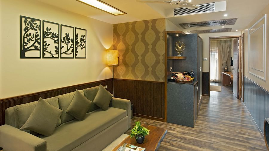 Image of Coral Suite at Coral Reef Hotel and Spa in Port Blair featuring a three-seater sofa with a wooden coffee table and a lamp in the corner as the shelf displays showpieces and snacks and the door leads to the bedroom