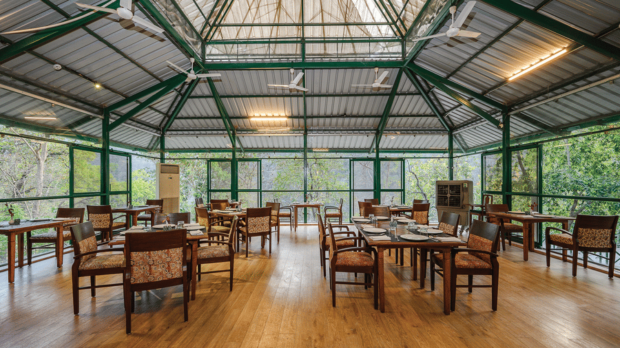 Spacious indoor dining room with a high glass ceiling and large windows that offer views of the surrounding trees at Neemrana’s Glasshouse on The Ganges featuring wooden floors and multiple dining tables for a light and airy mealtime experience