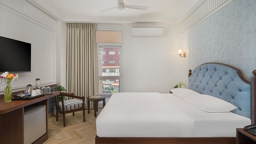 A spacious bed with a wall-mounted television above a wooden table, next to a large window with the curtains pulled to the side | Hotel Ganga Maiya