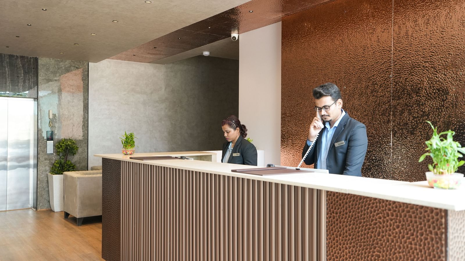 Two receptionists at a modern front desk, one on the phone and the other working on a computer - TGI Livancia Hillside, JC Nagar, Mysore