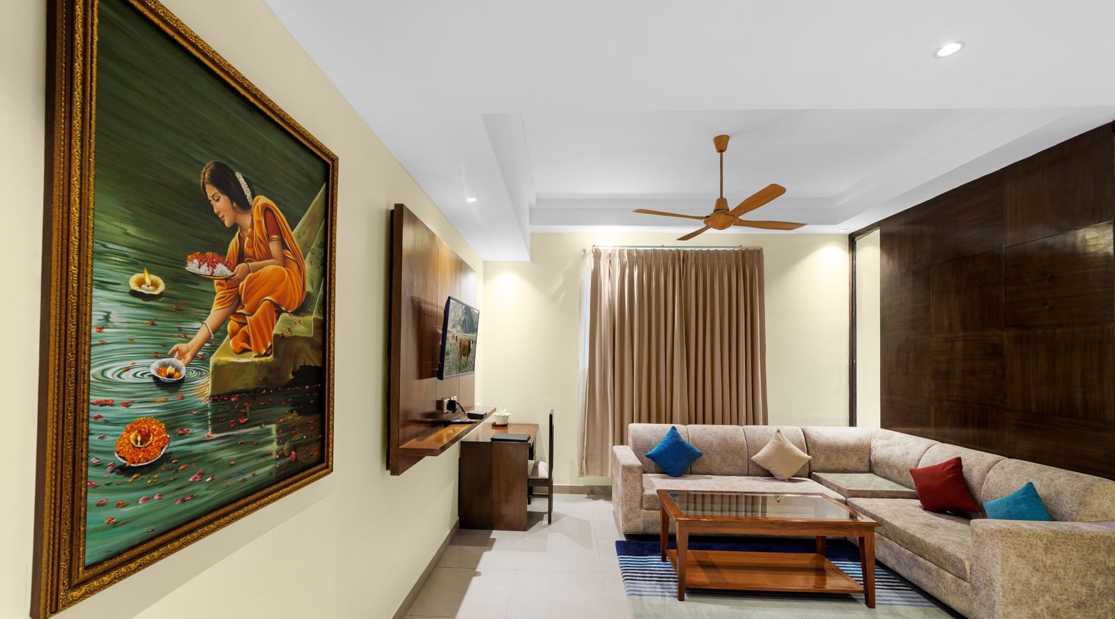 Living area at Inde Reva Resorts Rishhikesh with artwork on the wall, ceiling fan, and beige L-shaped sofa near curtains.