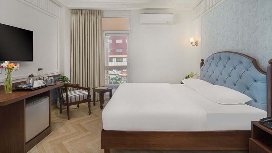 A spacious bed with a wall-mounted television above a wooden table, next to a large window with the curtains pulled to the side | Hotel Ganga Maiya