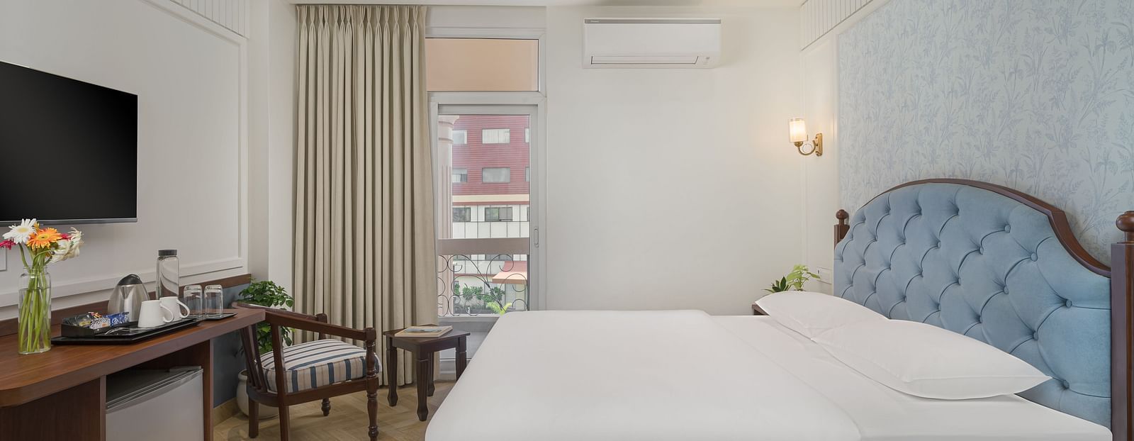 A spacious bed with a wall-mounted television above a wooden table, next to a large window with the curtains pulled to the side | Hotel Ganga Maiya