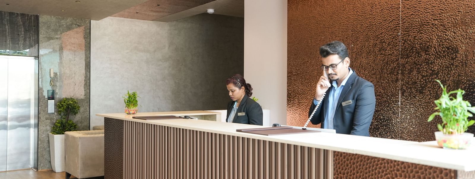 Two receptionists at a modern front desk, one on the phone and the other working on a computer - TGI Livancia Hillside, JC Nagar, Mysore