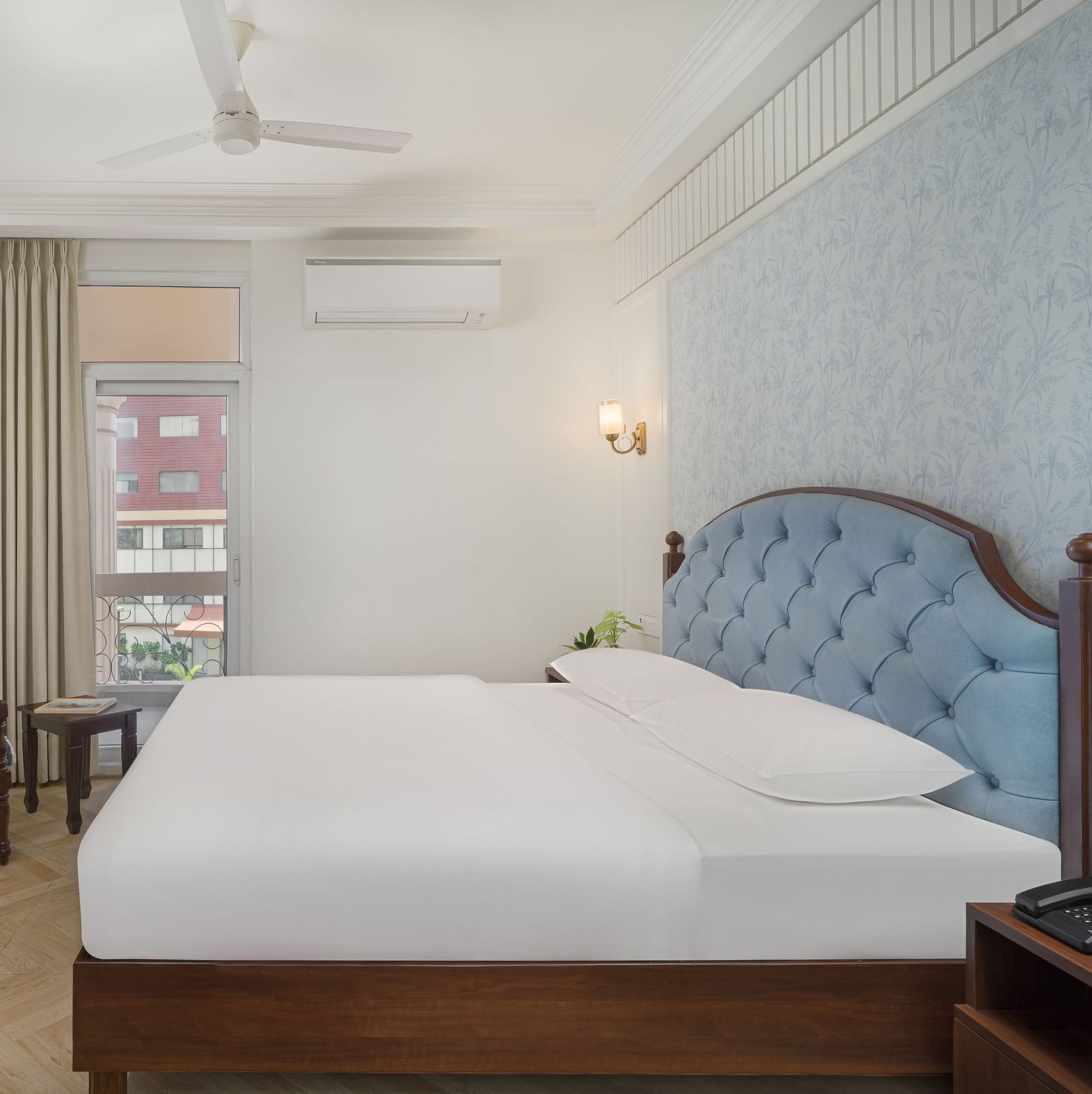 A spacious bed with a wall-mounted television above a wooden table, next to a large window with the curtains pulled to the side | Hotel Ganga Maiya