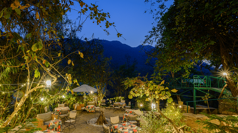 A beautifully lit outdoor dining area at dusk, surrounded by lush greenery with cosy tables, warm ambient lighting, and a backdrop of majestic mountains under the evening sky.