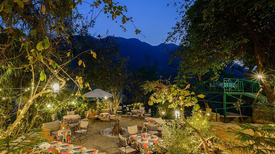 A beautifully lit outdoor dining area at dusk, surrounded by lush greenery with cosy tables, warm ambient lighting, and a backdrop of majestic mountains under the evening sky.