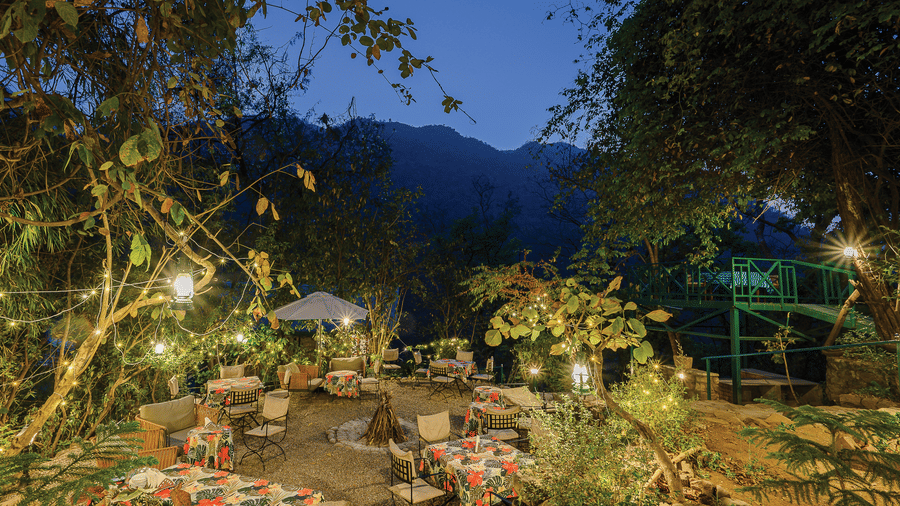 Atmospheric evening garden scene with dining tables and chairs illuminated by warm yellow lights under the forest canopy at Neemrana’s Glasshouse on The Ganges providing a magical outdoor setting for guests to enjoy their evening meals in the hills