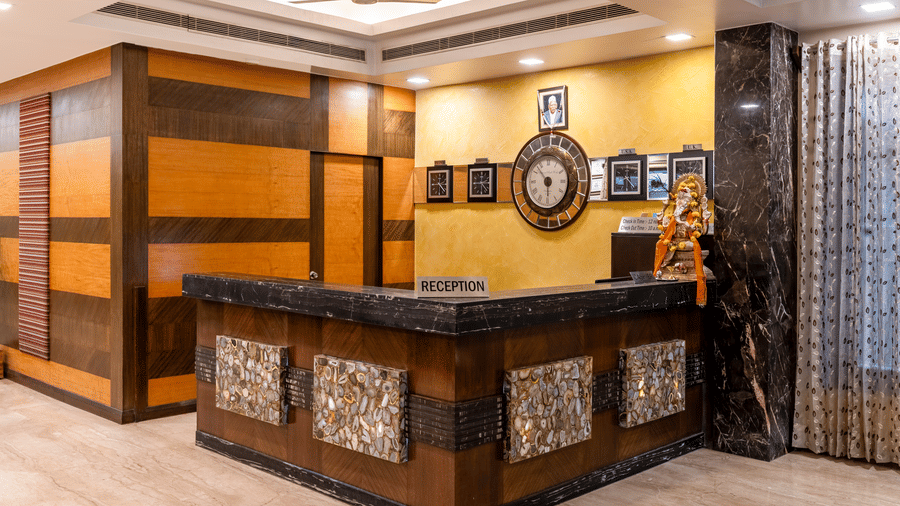 A close-up shot of the hotel reception desk featuring intricate wooden counter, a decorative analogue clock mounted on the wall, and warm overhead lighting - Amantra Comfort Hotel