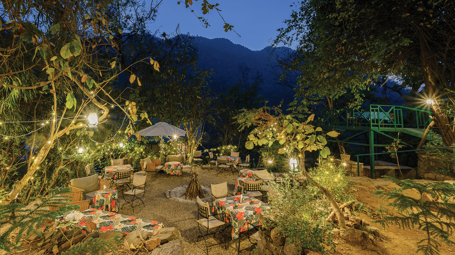 Atmospheric evening garden scene with dining tables and chairs illuminated by warm yellow lights under the forest canopy at Neemrana’s Glasshouse on The Ganges providing a magical outdoor setting for guests to enjoy their evening meals in the hills