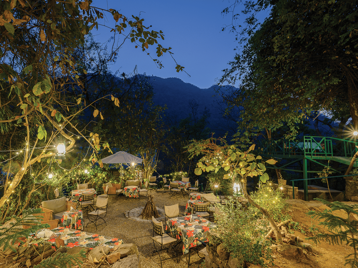 Atmospheric evening garden scene with dining tables and chairs illuminated by warm yellow lights under the forest canopy at Neemrana’s Glasshouse on The Ganges providing a magical outdoor setting for guests to enjoy their evening meals in the hills.